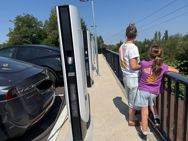 A Tesla Supercharger station in France featuring multiple charging stalls, a Tesla Model X, and adjacent buildings with shutters.