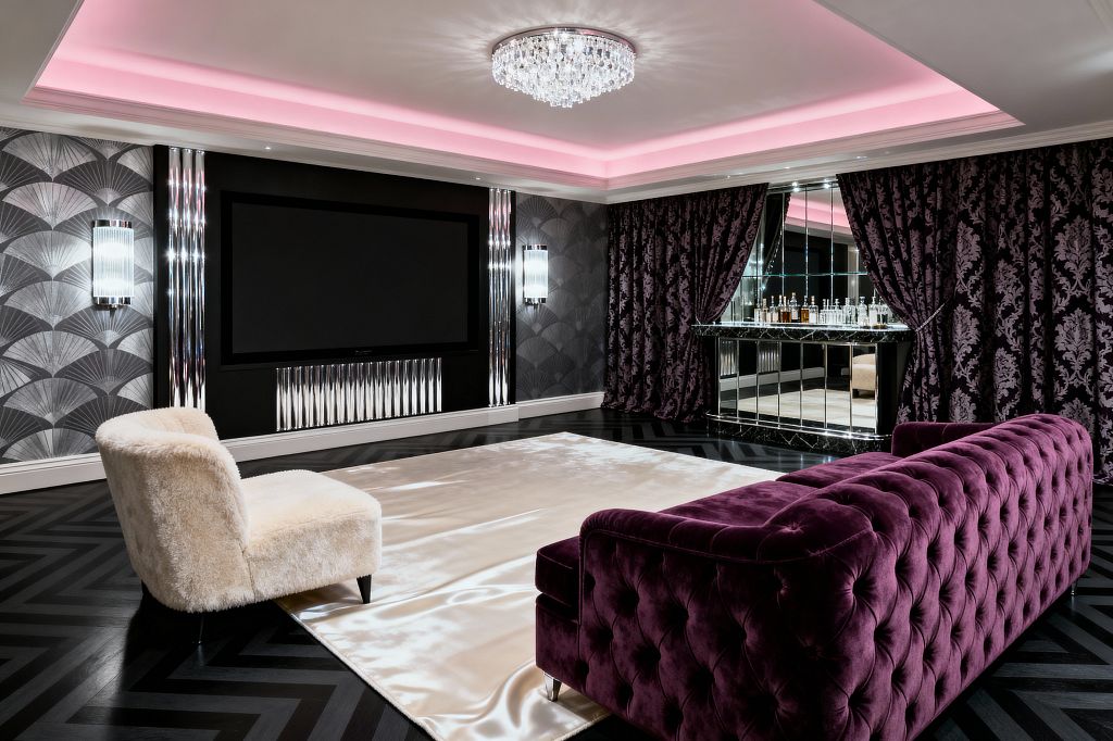 A glamorous media room pairs a deep plum tufted velvet sofa with an ivory faux‑fur lounge chair on a champagne satin rug over ebonized herringbone wood floors. A large black‑framed screen anchors an Art Deco wall with silver fan wallpaper, fluted chrome panels, and prismatic glass sconces, while a mirrored bar gleams behind purple damask curtains; a crystal ceiling light and pink cove lighting wash the tray ceiling, creating a dramatic vintage‑inspired atmosphere.