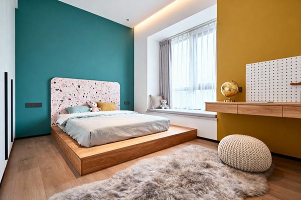 A modern children’s bedroom with a low light-oak platform bed, mint bedding, and a rounded pink terrazzo-pattern headboard set against a teal wall. A mustard accent wall features a floating wood desk with a white pegboard and gold globe, while a large window with sheer curtains and a cushioned bench brightens the space; a knitted cream pouf and fluffy rug soften the oak floor, and concealed cove lighting adds a warm glow.
