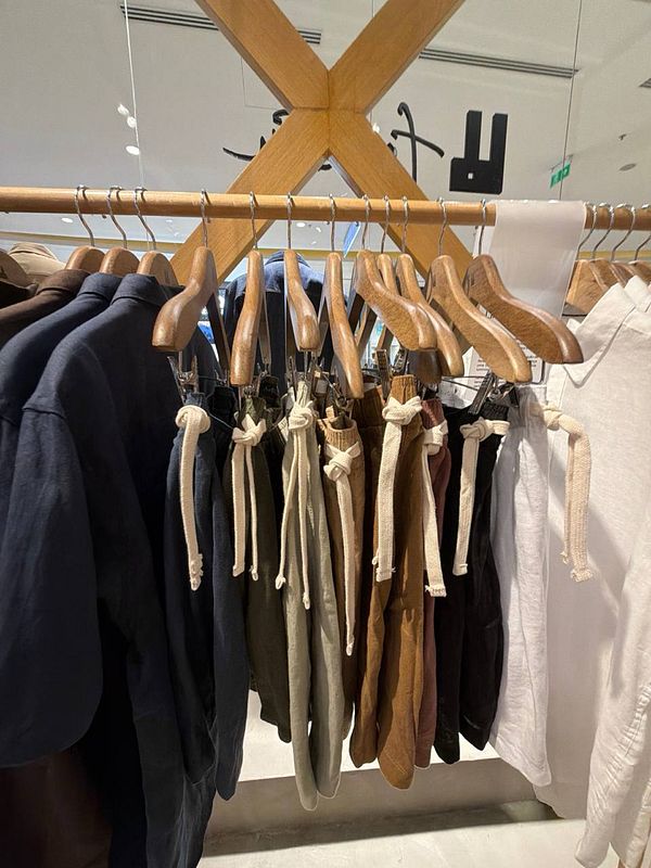 A man in a casual outfit composed of a sky-blue shirt, beige linen shorts, and white sneakers stands outdoors on a concrete ledge with greenery in the background.