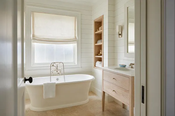 A serene coastal-style master bathroom with a freestanding white soaking tub centered beneath a large window with a soft Roman shade. White shiplap walls, a light wood vanity with brass hardware, open shelving, and pale stone flooring create a calm, airy space.
