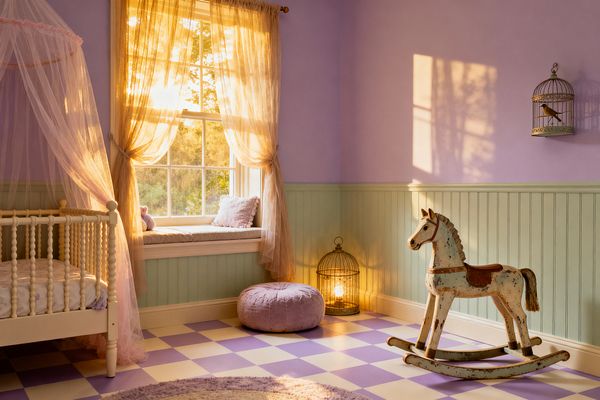 A pastel kids’ room features lavender walls above pale mint beadboard and a lilac-and-cream checkerboard floor. A vintage distressed wooden rocking horse stands near a large window with gauzy champagne curtains, a cushioned window seat with pillows, and a round lilac pouf; to the left is a white spindle crib draped with a blush tulle canopy, while a brass-tone birdcage lamp and wall birdcage provide warm, whimsical accents.
