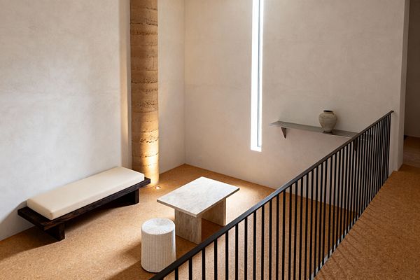 A minimalist living room with warm plaster walls features a cork floor, a rectangular travertine coffee table, and a low dark-stained wood bench topped with a cream cushion. A white woven cylindrical stool sits nearby; a narrow vertical window supplies daylight; a textured earthen column is lit from a floor uplight; a slim metal floating shelf displays a single ceramic vase; and a black metal balustrade lines the mezzanine edge, creating a serene, uncluttered atmosphere.