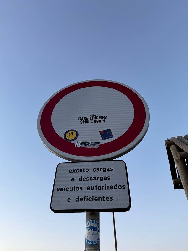 A street sign with a playful message and additional stickers is prominently displayed against a clear sky.