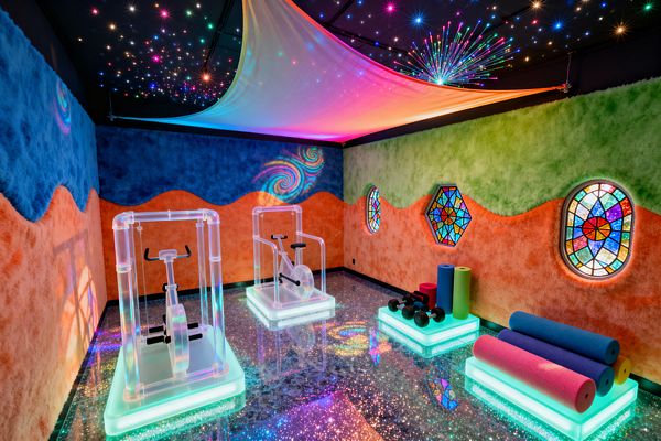 Home gym featuring two transparent acrylic stationary bikes on glowing LED plinths, with adjacent illuminated platforms holding colorful foam rollers and black dumbbells. A glitter-speckled epoxy floor reflects wavy blue, lime, and tangerine textured walls punctuated by backlit stained-glass windows, while a black fiber‑optic star ceiling and suspended rainbow fabric canopy flood the room with prismatic light and subtle swirl projections.