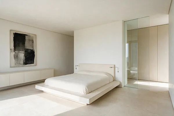 A minimalist master bedroom with a low white platform bed centered against a plain white wall, a large abstract black-and-white painting on the left, and a long встроенный-style storage cabinet beneath it. On the right, a glass partition reveals a built-in wardrobe with pale wood panels. The room uses soft neutral tones, clean lines, and abundant daylight on a light stone floor.