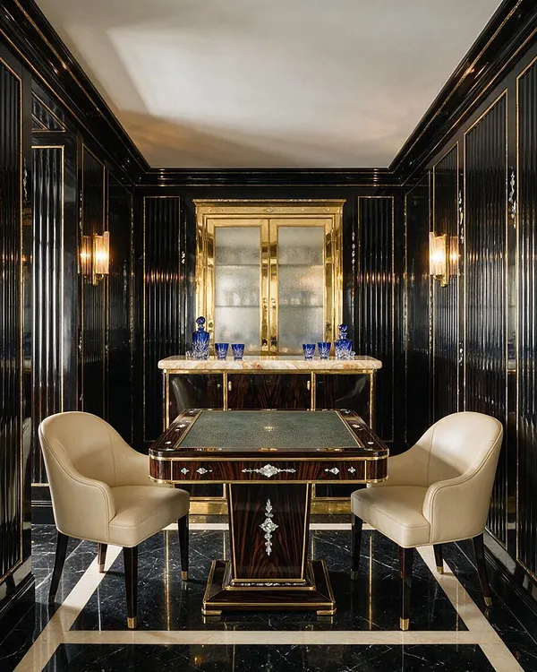 A narrow Art Deco game room with glossy black paneled walls trimmed in gold, a black marble floor with cream border lines, and a central card table. Two cream upholstered chairs flank the table, while a gold-and-glass bar cabinet stands at the back with blue glass decanters and tumblers.