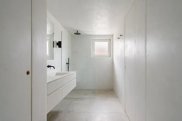 A minimalist basement bathroom with smooth white walls, a floating double-sink vanity, matte black fixtures, and a small frosted window bringing in natural light. Large pale stone-look floor tiles and a frameless mirror keep the space clean and simple.
