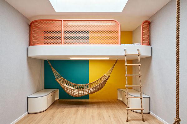 A contemporary kids' room with a loft bed edged in rounded white forms and a coral perforated metal guardrail beneath a skylight. Below, a natural-rope hammock hangs before a teal and mustard color-block wall, flanked by two curved white storage benches; a rope ladder drops from the loft and a separate climbing rope hangs to the right, with light oak flooring, pale gray walls, and a linear LED under the loft for illumination.