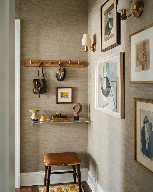 A narrow eclectic entryway with textured beige wallpaper, framed abstract art, and brass wall sconces. A wooden peg rail, clear floating shelf with small decorative objects, and a leather-topped stool add warmth and character.
