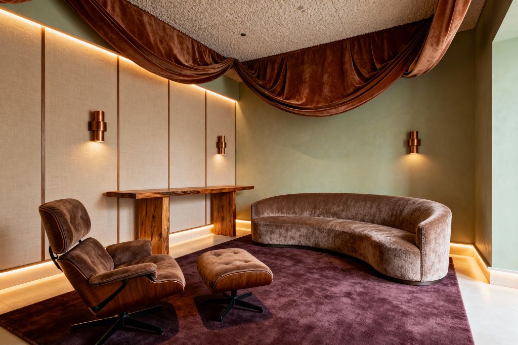 A transitional media room features a curved taupe velvet sofa and a brown lounge chair with matching ottoman on a deep plum rug. Sage plaster walls and a linen-clad panel wall with wood trim are washed with warm LED cove lighting and copper sconces. A live-edge wood console anchors the wall, and draped rust velvet runs across the ceiling line, softening the textured surface and enhancing acoustics to create a cocooned, cinema-ready atmosphere.