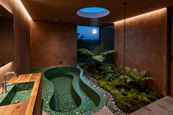 A biophilic bathroom features a sculptural green terrazzo soaking tub and a matching integrated sink set into a teak vanity. Earthy terracotta plaster walls and warm cove lighting frame an indoor garden of moss, ferns, and river pebbles beside a floor-to-ceiling corner window showing the night sky and full moon; a circular skylight above creates a gentle rain cascade, echoed by a hanging rain chain and basin for a tranquil, nature-connected experience.