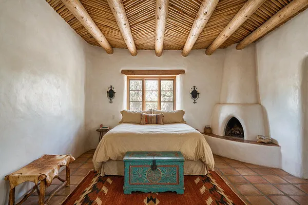 A Southwestern-style guest bedroom with white adobe walls, a woven wood ceiling with exposed beams, and a centered bed dressed in soft beige bedding. Terracotta tile floors, a turquoise painted chest at the foot of the bed, a cowhide bench, and a small kiva-style fireplace create a warm rustic atmosphere.