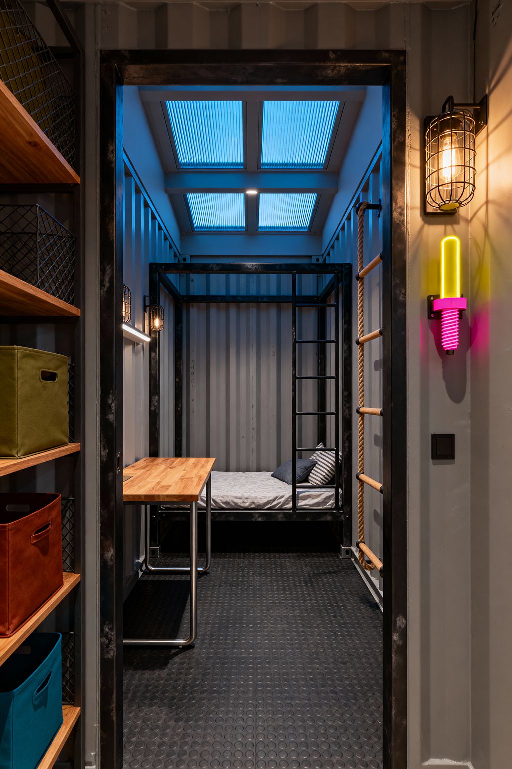 A compact kids’ room with corrugated metal walls shows a black steel bed frame with an integrated ladder, a small desk with a light wood butcher-block top on metal legs, and black rubber coin flooring. Blue-tinted polycarbonate skylight panels brighten the space, highlighting wire-caged wall lights, a neon pink and yellow sconce, and a wall-mounted rope ladder with wooden rungs. Open shelving on the left holds wire baskets and colorful fabric storage bins.