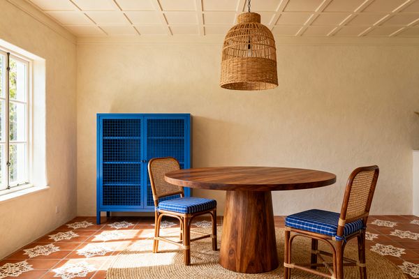 A sunlit French Country dining room with cream plaster walls, a coffered ceiling, and terracotta floor tiles stenciled with white motifs. At center sits a round solid-wood pedestal table on a jute rug, paired with two cane-back rattan chairs upholstered in blue striped fabric. A woven rattan dome pendant hangs above, and a cobalt-blue metal cabinet with mesh doors stands against the wall beside a multi-pane window flooding the space with light.
