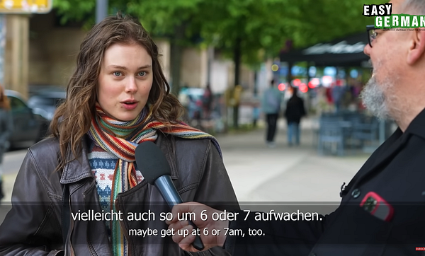 A young woman being interviewed on the street as part of an Easy German video, discussing waking up times with German and English subtitles.