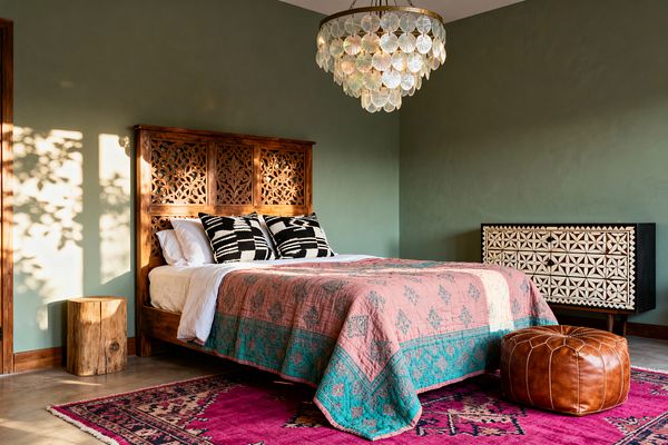 A Bohemian bedroom with sage green walls features a carved wooden bed with white linens, two black-and-ivory patterned pillows, and a blush quilt edged in teal. A vivid magenta rug with geometric motifs covers the floor, paired with a cognac leather pouf and a raw log nightstand. To the right stands a black dresser with cream bone-inlay patterns, while a brass-framed capiz shell chandelier glows as afternoon light casts leafy shadows on the walls.