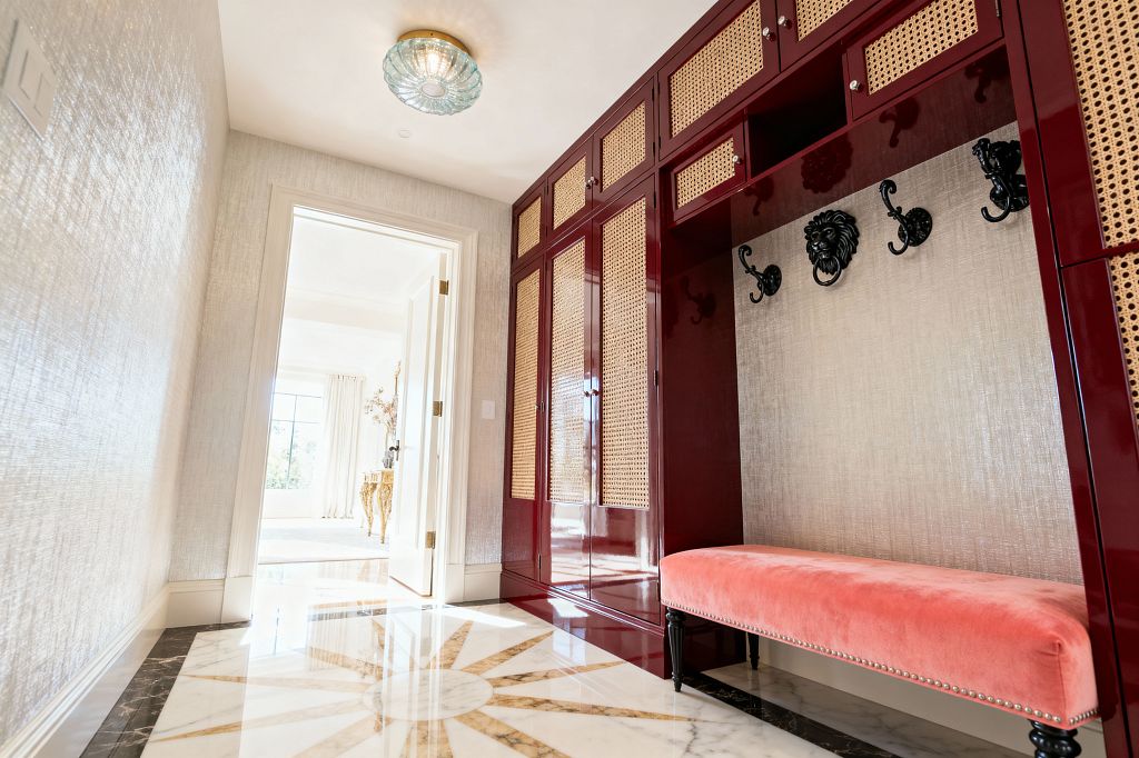 A glam mudroom features glossy burgundy built-in lockers with cane-panel doors and a coral velvet bench with nailhead trim. Black sculptural hooks, including a central lion-head design, mount on champagne metallic wallcovering. The polished marble floor has a sunburst inlay framed by a dark border, and a brass-based ribbed glass flush-mount light crowns the ceiling near an open doorway to a bright adjoining room.