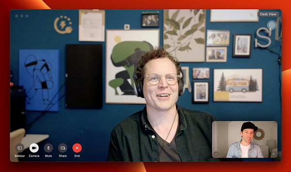 A video call featuring two individuals, one smiling and wearing headphones, with a colorful background of artwork and photos.