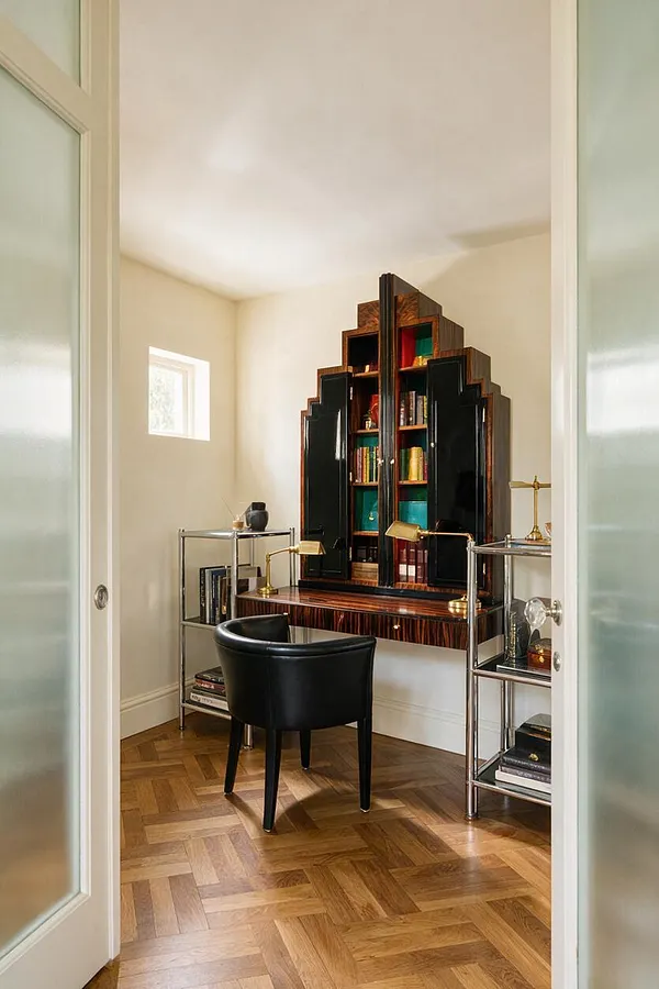 A small Art Deco-inspired backyard room with cream walls, a warm herringbone wood floor, and a dramatic stepped black-and-wood cabinet centered on the back wall. A black upholstered chair sits in front of a slim desk, flanked by chrome shelving units with brass accents and decorative objects, creating a polished, elegant atmosphere.