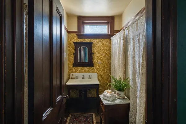 A narrow Victorian-style powder room with dark wood trim, yellow floral wallpaper, and a small high window above a wall-mounted sink. Lace shower curtains line the right side, while a small cabinet with folded towels and a fern adds a homey touch.