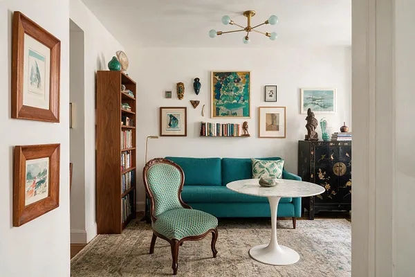 A bright eclectic living room with a teal sofa, patterned accent chair, and white round pedestal table centered on a textured rug. The walls are filled with mixed artwork, a tall wood bookshelf stands to the left, and a black lacquer cabinet with decorative objects adds contrast on the right.