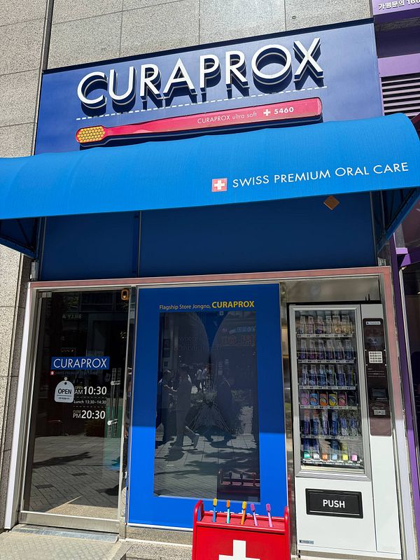 A modern Curaprox flagship store interior featuring dental care products on display with a large screen showing a model's smiling face.