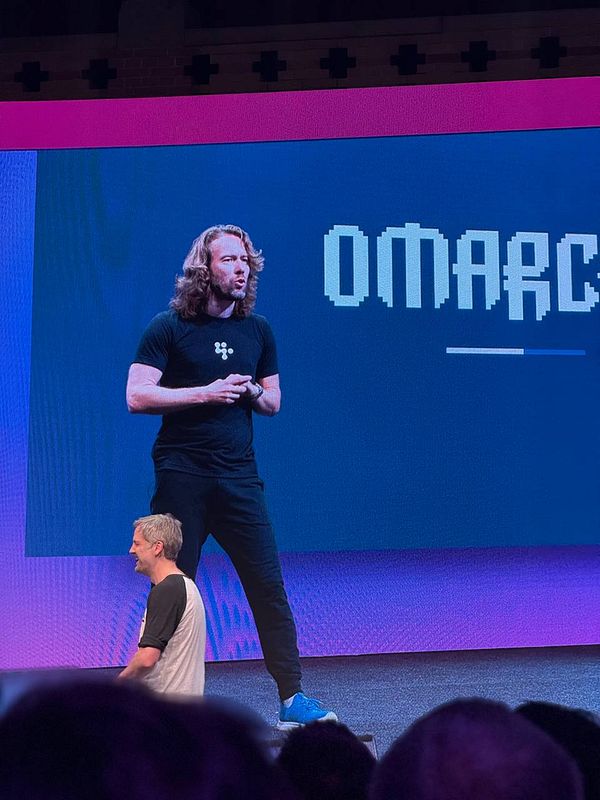 A speaker presents on stage at a conference, with a large screen displaying text behind them.