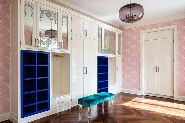 An elegant mudroom with glossy ivory built-in cabinetry featuring etched mirrored doors and polished nickel pulls, flanked by bold cobalt blue cubbies. Pink Art Deco fan wallpaper lines the walls, a teal velvet bench with clear acrylic legs sits on herringbone walnut flooring, and a smoked-plum glass pendant with brass details hangs overhead, creating a glamorous yet organized entry space.