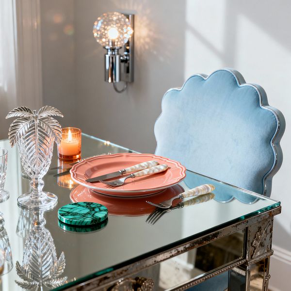 Close-up of a glamorous dining setting: a mirrored table reflects a powder-blue velvet chair with a scalloped back and silver piping. On the tabletop are a coral scalloped plate with pearl-handled knife and fork, a small amber candle, a cut-crystal palm-shaped vase, a clear tumbler, and a green malachite-pattern coaster. A polished-chrome wall sconce with a faceted globe throws sparkling light across soft white walls.