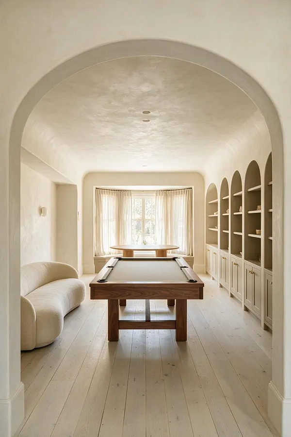 A serene game room with a central wooden pool table, a curved upholstered sofa on the left, and built-in arched shelving with cabinets on the right. Soft cream walls, pale wood flooring, sheer curtains, and a rounded archway create a warm Organic Modern atmosphere.