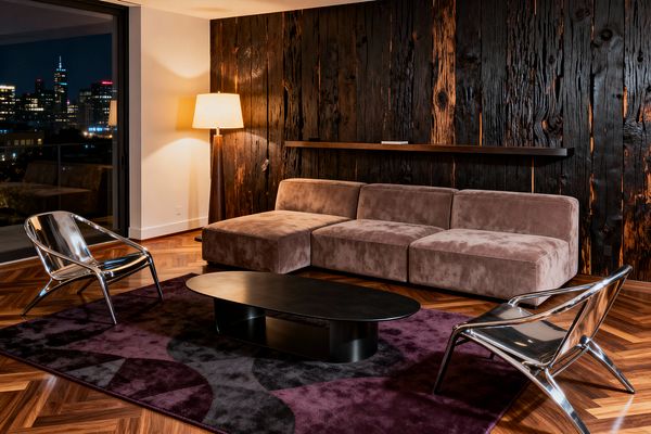 Contemporary living room with a charred wood plank feature wall and slim floating shelf, a low taupe velvet sectional, and two polished chrome lounge chairs. A matte black oval coffee table rests on a deep plum and charcoal rug. A warm floor lamp highlights the walnut herringbone floor, while a floor-to-ceiling window reveals a nighttime city skyline, adding reflections and a soft ambient glow to the space.