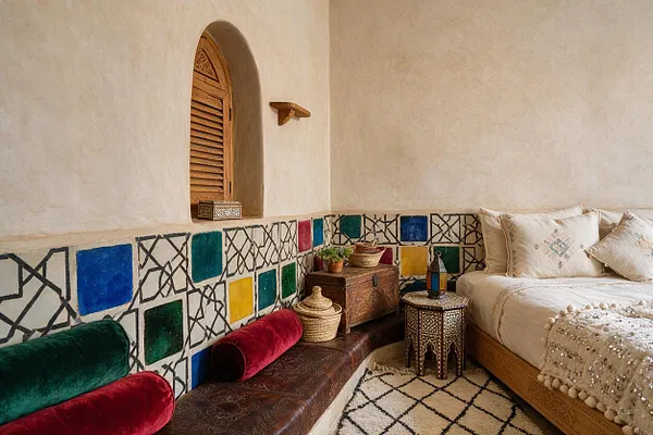 A Moroccan-style master bedroom with cream plaster walls, an arched wooden shutter window, and a low built-in bench lined with leather and velvet bolsters. The lower wall is painted with colorful square tile-like motifs in blue, green, yellow, and pink, while carved wood furniture, woven baskets, brass lanterns, and a cream bed with patterned pillows create a warm, layered look.