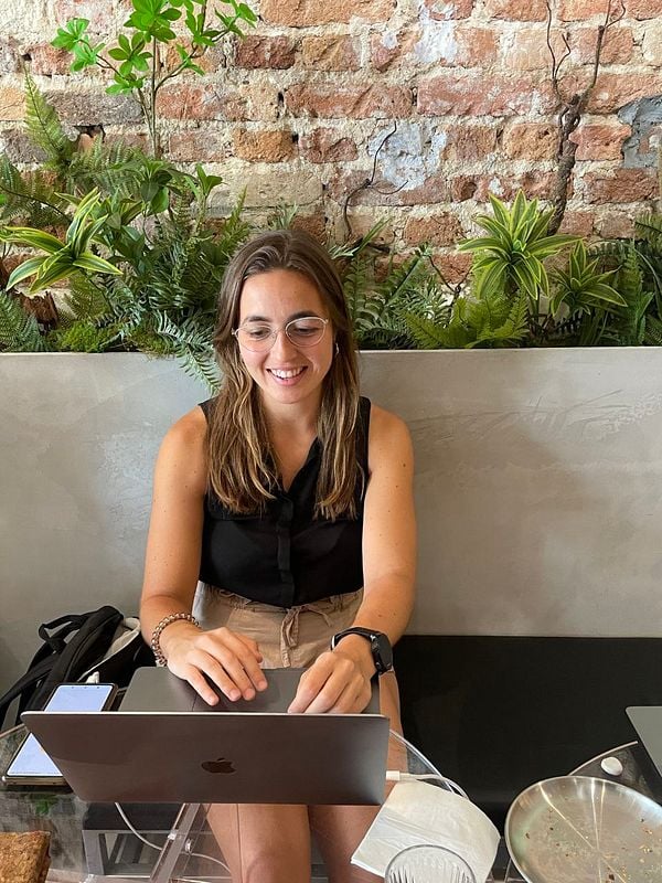 A woman is sitting at a table in a cafe, working on her laptop with a smile.