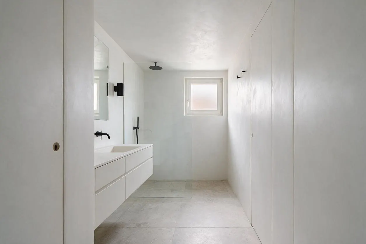 A minimalist basement bathroom with smooth white walls, a floating double-sink vanity, matte black fixtures, and a small frosted window bringing in natural light. Large pale stone-look floor tiles and a frameless mirror keep the space clean and simple.