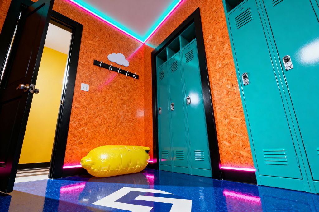A vibrant mudroom features teal metal lockers set in black-framed niches against orange OSB walls. A glossy electric-blue floor with a white graphic reflects neon pink base lighting and a cyan strip along the ceiling. A cloud-shaped plaque tops a black hook rail, and a yellow inflatable lemon serves as a playful seat. A black door opens to a bright yellow adjoining room. Chrome locker latches and bold contrasts create a Pop Art, utility-meets-fun atmosphere.