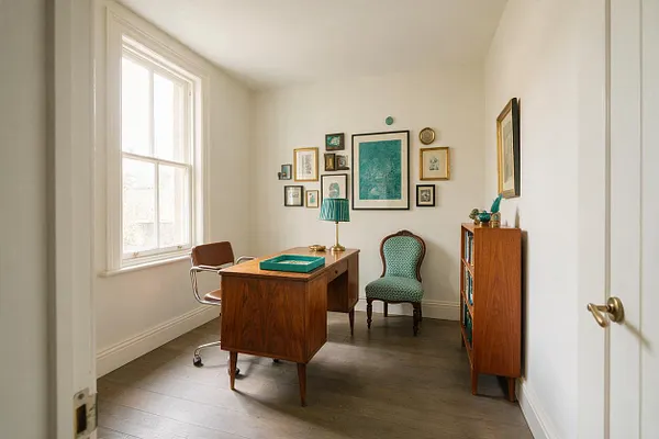 A compact eclectic home office with white walls, a large window on the left, and dark wood floors. The room features a mid-century wooden desk with a teal tray, a turquoise upholstered chair, a framed gallery wall with teal artwork, a brass lamp, and a small wooden bookshelf on the right.
