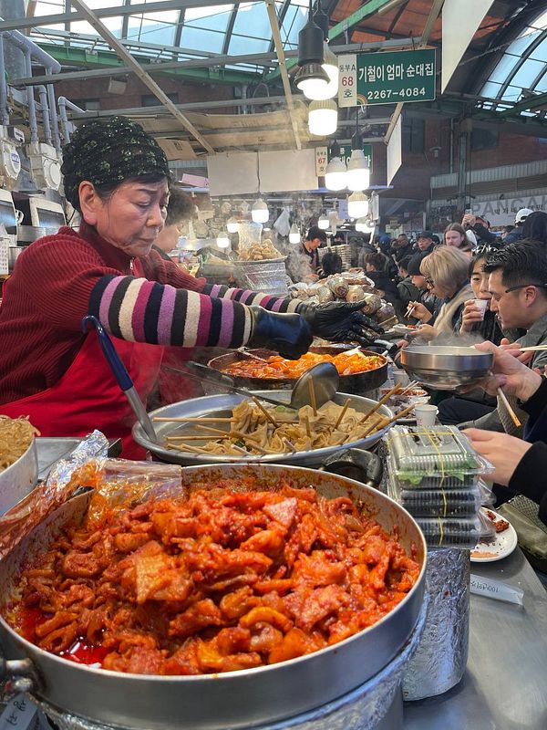 A bustling market scene featuring a vendor serving various Korean street foods.