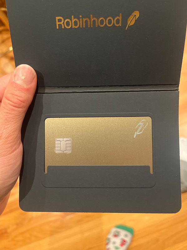 A hand holding an opened black presentation cardholder with a gold Robinhood credit card inside.