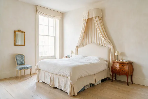 A serene master bedroom in French Country style with a cream upholstered bed draped in sheer canopy fabric, pale walls, and a large tall window bringing in daylight. The room includes a carved wooden bedside table, a small upholstered chair with blue checks, a gold-framed wall mirror, and light wood floors.