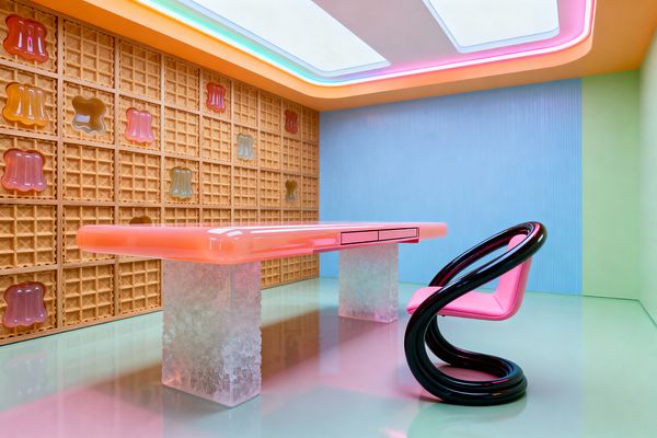 A playful home office features a waffle-patterned wall inset with glossy, jelly-like forms in pink, amber, and green. Centered is a rounded bubblegum-pink desk with two slim drawers on frosted resin block legs, paired with a sculptural black loop chair with a pink seat; the mint-gloss floor reflects pastel light from a ceiling of backlit panels and neon coves, while powder-blue and pale-green walls complete the candy-inspired palette.