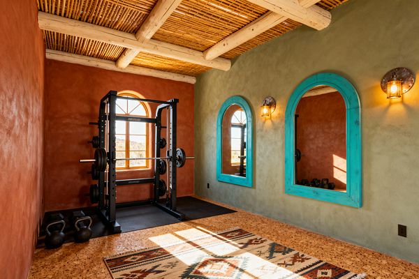 A Southwestern-style home gym with a textured terracotta wall facing a sage green wall that holds two arched mirrors framed in turquoise. A black steel power rack with barbell and plates sits on black rubber mats beside kettlebells. Warm caged wall sconces flank the mirrors. The ceiling features pale log beams with woven reed latillas, and the floor appears cork with a geometric kilim rug. An arched wood window lets in natural light and shows desert views.