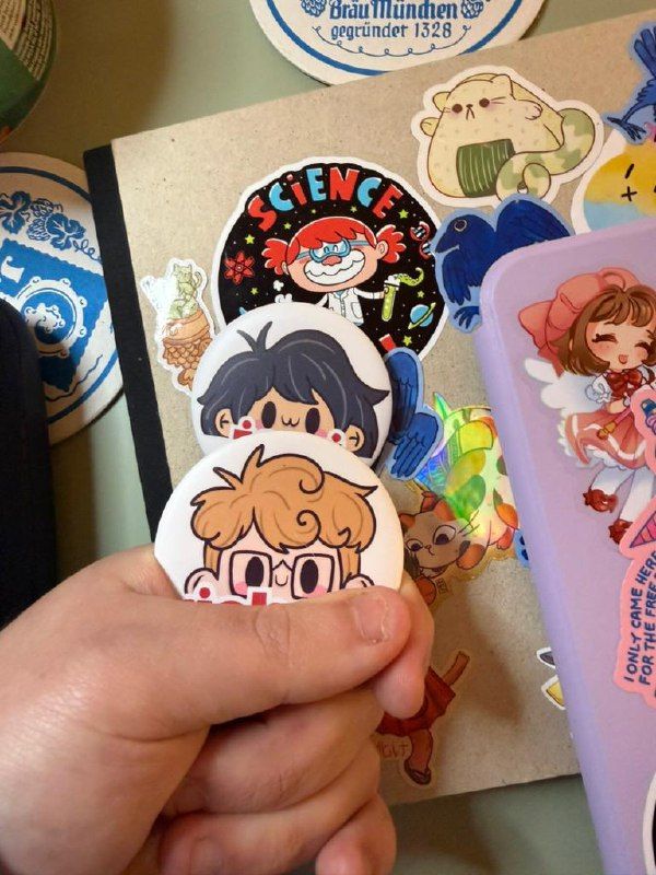 A hand holds three cartoon-style name pins featuring characters with distinct hairstyles and expressions.