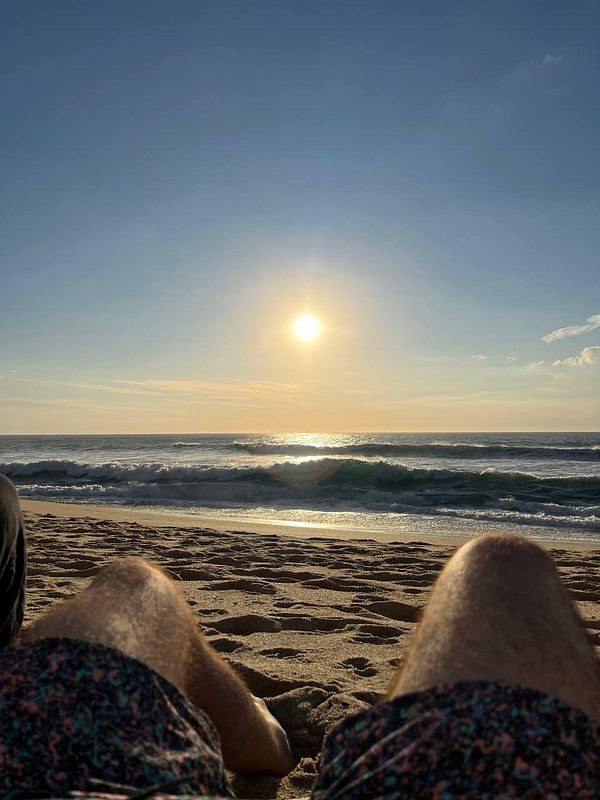 A serene beach scene at sunset with a focus on two pairs of legs resting on the sand.