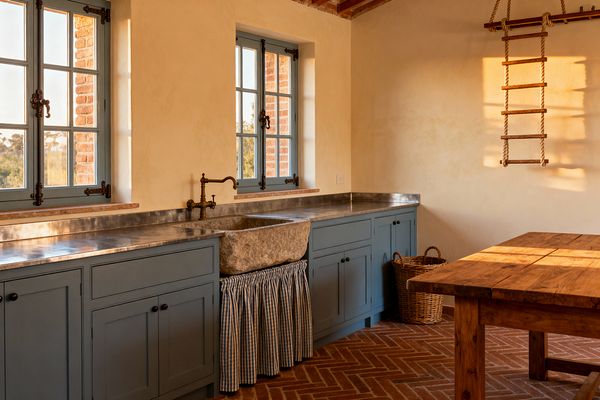 A rustic laundry room shows dusty blue Shaker cabinets spanning one wall beneath three casement windows, topped with a zinc work surface and a low metal backsplash. Centered is a carved stone apron-front sink with a wall-mounted brass faucet and a blue-and-white gingham skirt, surrounded by warm plaster walls, herringbone terracotta flooring, an exposed wood-and-brick ceiling, a wicker basket, a rope ladder-style drying rack, and a chunky farmhouse table in golden light.