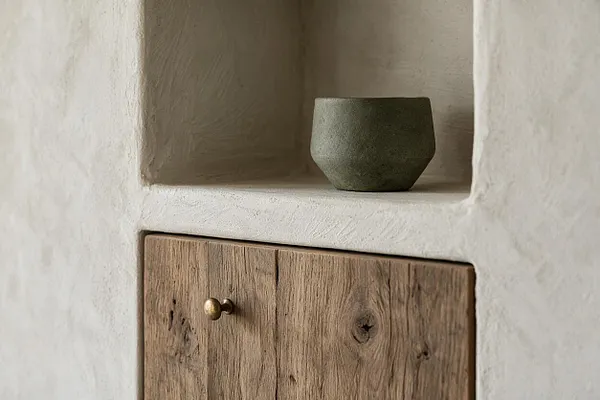 A Wabi-Sabi style wall detail with a softly rounded plaster niche holding a small matte ceramic vessel above a rustic wooden cabinet door with a simple brass knob. The off-white plaster, weathered wood grain, and muted olive-green bowl create a calm, handcrafted look.