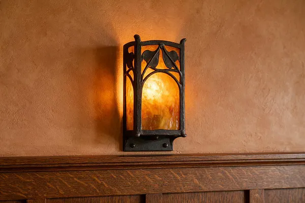 A Craftsman-style wall sconce mounted on a warm tan plaster wall above dark wood wainscoting. The fixture has a black metal frame with arched, decorative lines and glowing amber glass that casts a soft golden light.