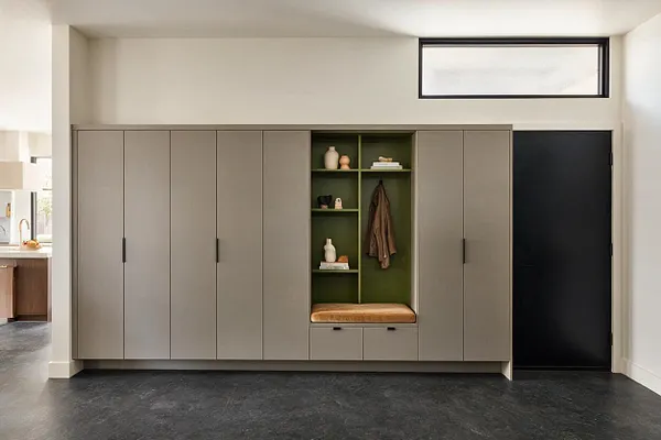 A modern entryway with a wall of tall taupe built-in cabinets and a central open niche in olive green. The niche includes shelves, a hanging rail with a coat, a cushioned bench, and small decor objects, with a black door and transom window nearby.