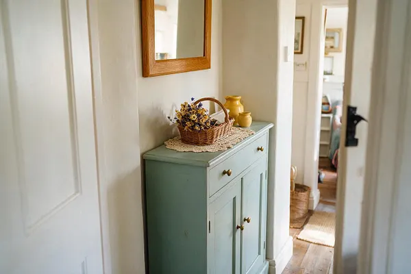 A narrow cottagecore entryway with a soft sage cabinet topped by a lace doily, wicker basket of dried flowers, and small yellow ceramic vessels. A wooden-framed mirror hangs above, and the warm wood floor and woven textures create a cozy, lived-in feel.