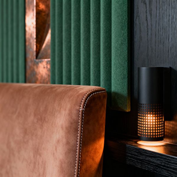 Close-up of a modern media room wall with vertical forest-green felt acoustic panels next to a black-stained oak ledge. In front sits a caramel suede upholstered backrest with white contrast stitching. A matte black perforated cylindrical wall sconce casts a warm amber glow across the wood and leather, while a narrow oxidized copper inlay adds a metallic accent, emphasizing texture, sound control, and low, cinematic lighting.